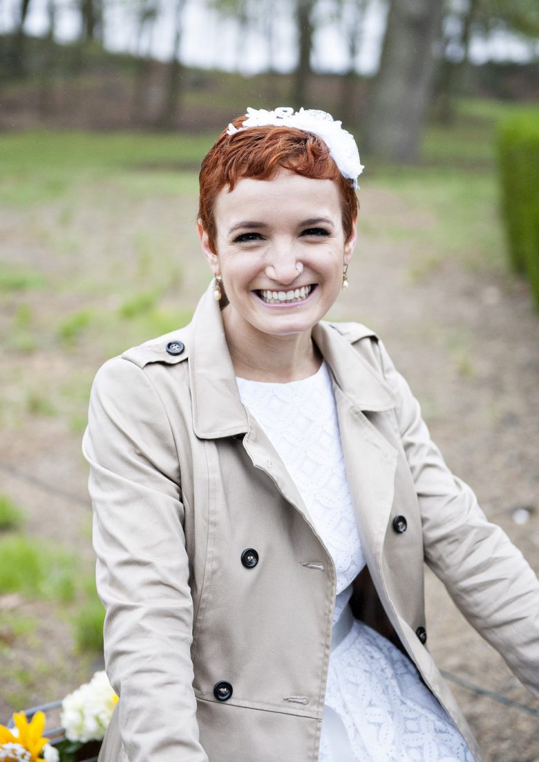 A woman in a white wedding dress and khaki trench coat wears her red hair in a pixie cut with floral headpiece and smiles