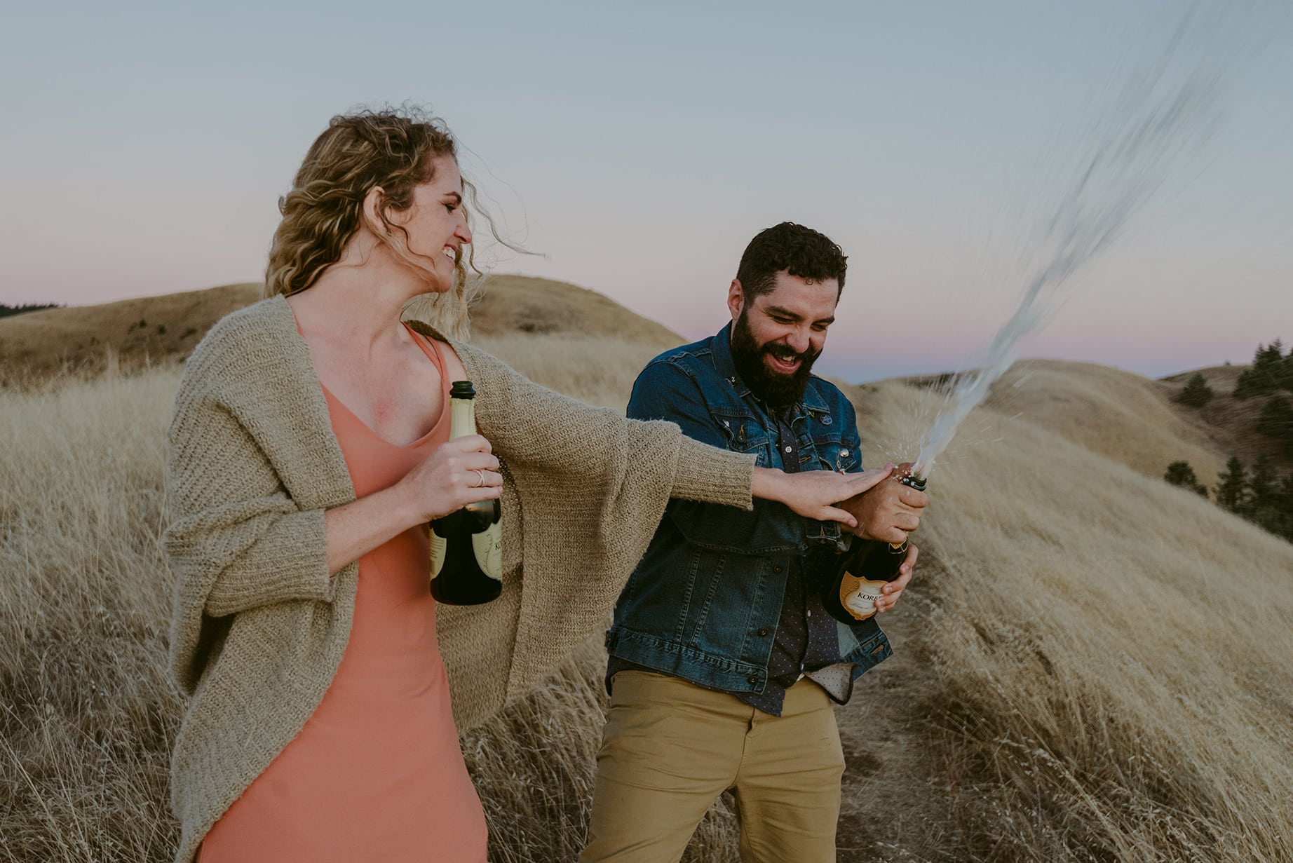 A Julie Pepin photo depicting a couple opening a bottle of champagne on grassy dunes. The champagne is shooting out of the bottle and the couple laughs
