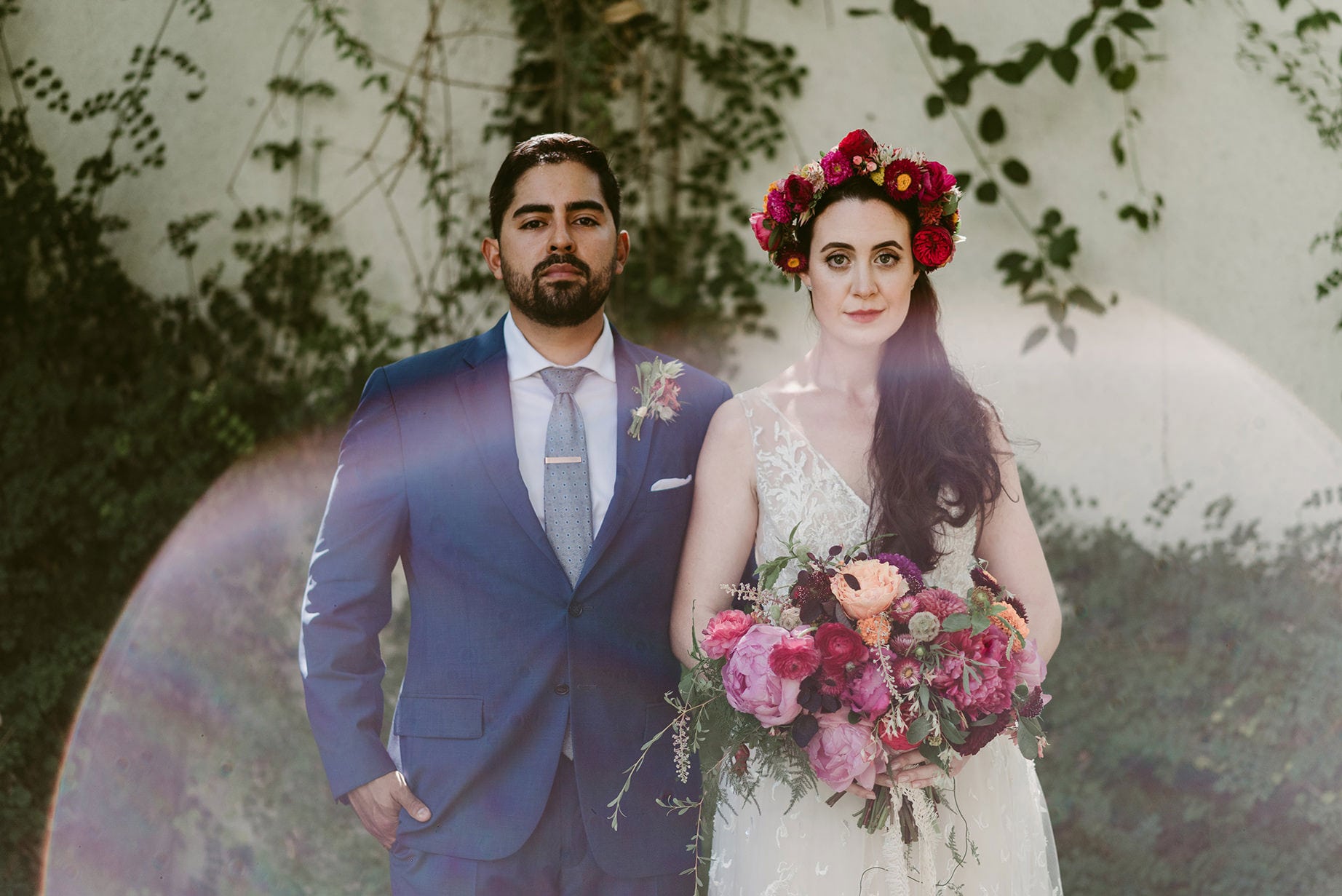 A Julie Pepin photo depicting a wedding couple. A groom wears a blue suit and tie, and a bride is in a white gown with a lace illustion neckline and a dark pink and yellow flower crown and bouquet.