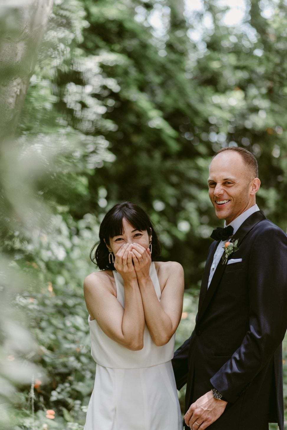 A Julie Pepin photo depicting a bride with shoulder length dark hair and bangs laughing into her hands, standing next to a groom in a black tux and bow tie, also laughing
