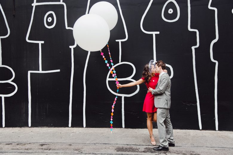 Stylish couple on a black and white wall with balloons, the bride wears a DIY birdcage veil and a red dress