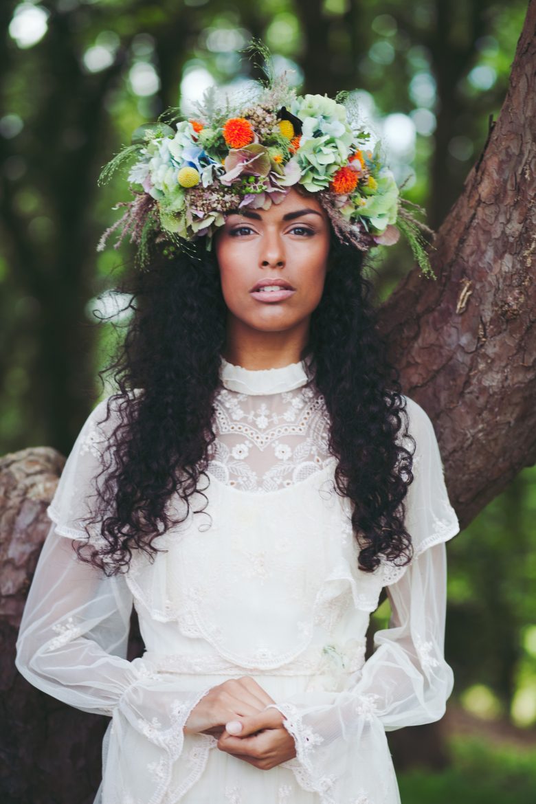 woman with long dark corkscrew curls and large bright flower crown, wearing a high-necked long-sleeved wedding dress