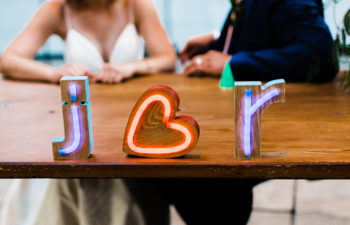 Neon block letters read "j heart r" on a wood table in front of a couple in wedding clothes