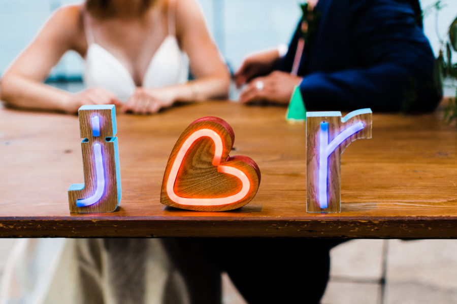 Neon block letters read "j heart r" on a wood table in front of a couple in wedding clothes