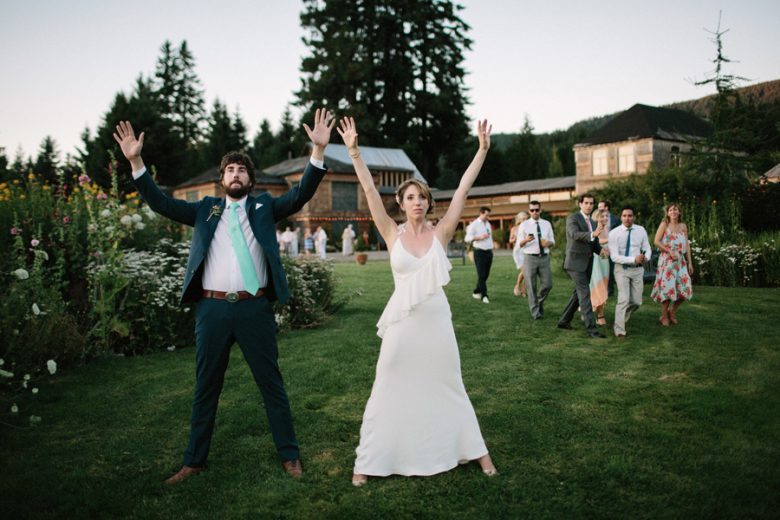 bride and groom outside a cabin facing camera with guests walking behind them