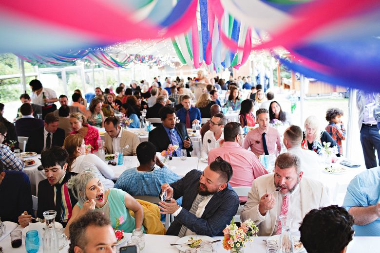Many people eating long white tables with colorful streamers | A Practical Wedding