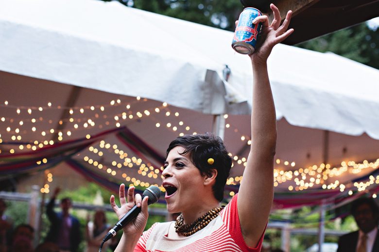 Woman with microphone holding up beer for a toast with string lights in the distance | A Practical Wedding