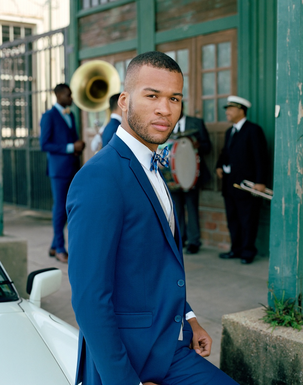 a man wearing a blue wedding suit and a plaid bowtie from generation tux stares into the camera while a new orleans jazz band stands behind him