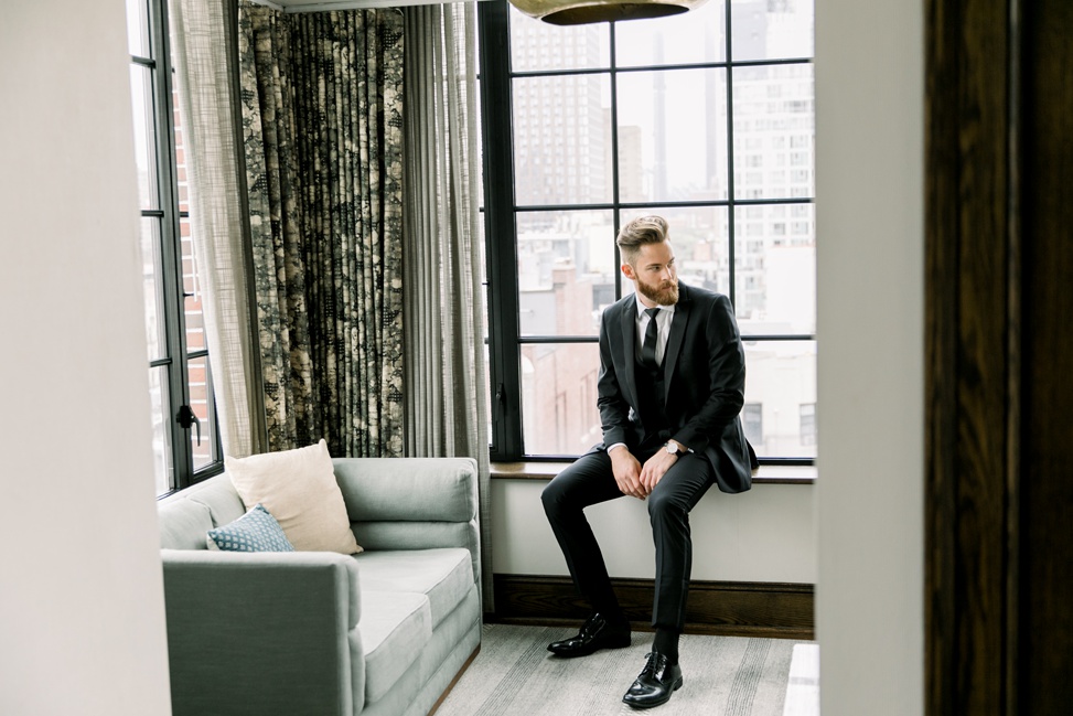 a man sits on a windowsill wearing a black wedding suit for men from generation tux