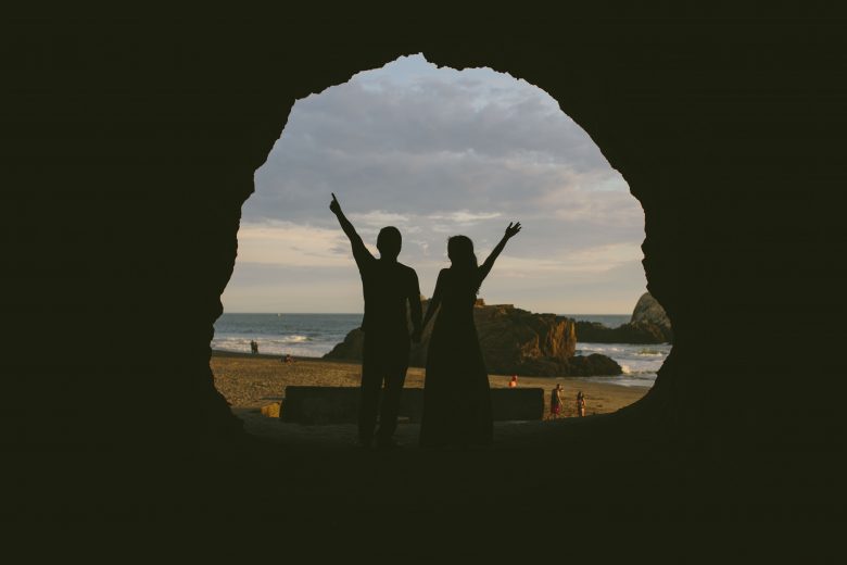 silhouette of woman and man standing in cave in front of the ocean