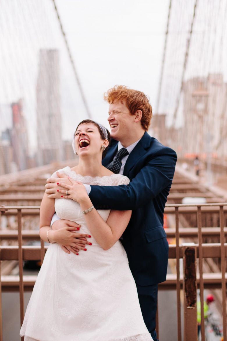 couple laughing on the brooklyn bridge wearing wedding outfits