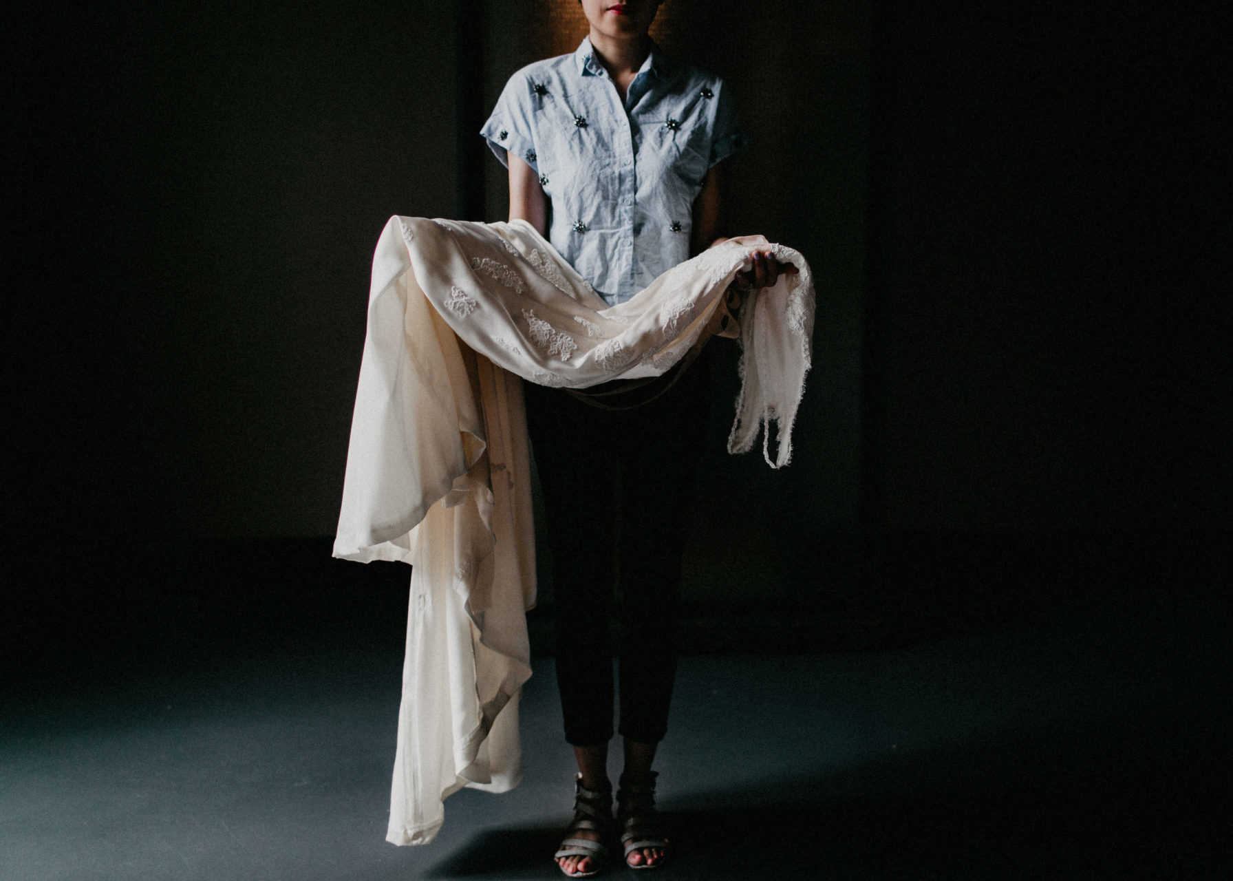A woman in an embellished short-sleeved chambray top holds a wedding dress draped over her arms in a dark, shadowy space in a photo by Shaw Photography Co.