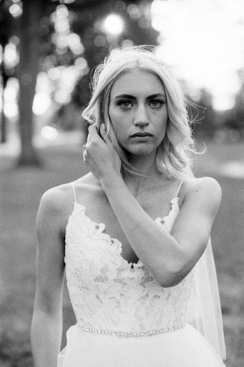 A bride in a spaghetti strap dress with a lace bodice brushes back her hair and looks intensely at you in a black and white photo by Shaw Photography Co.