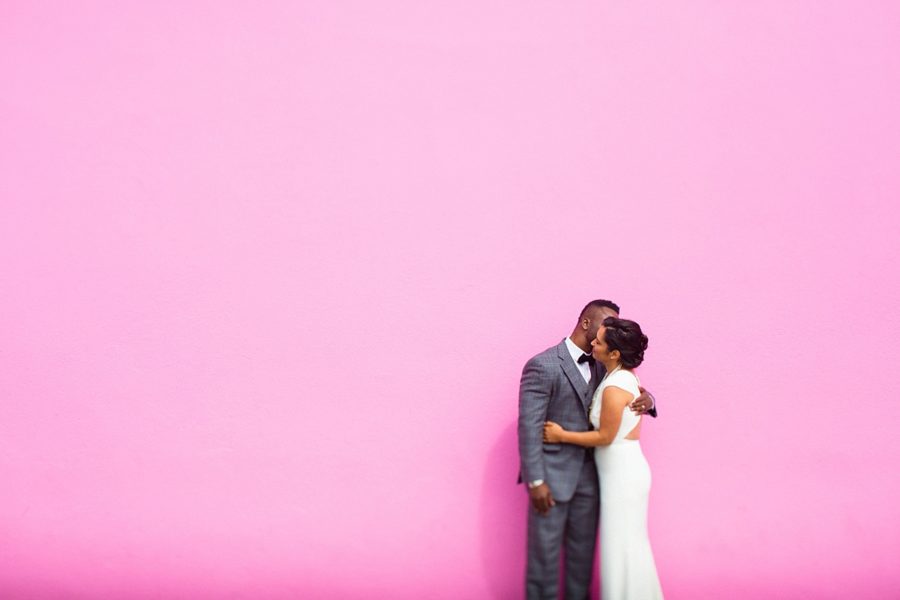 A couple in wedding clothes embraces in front of a pink wall