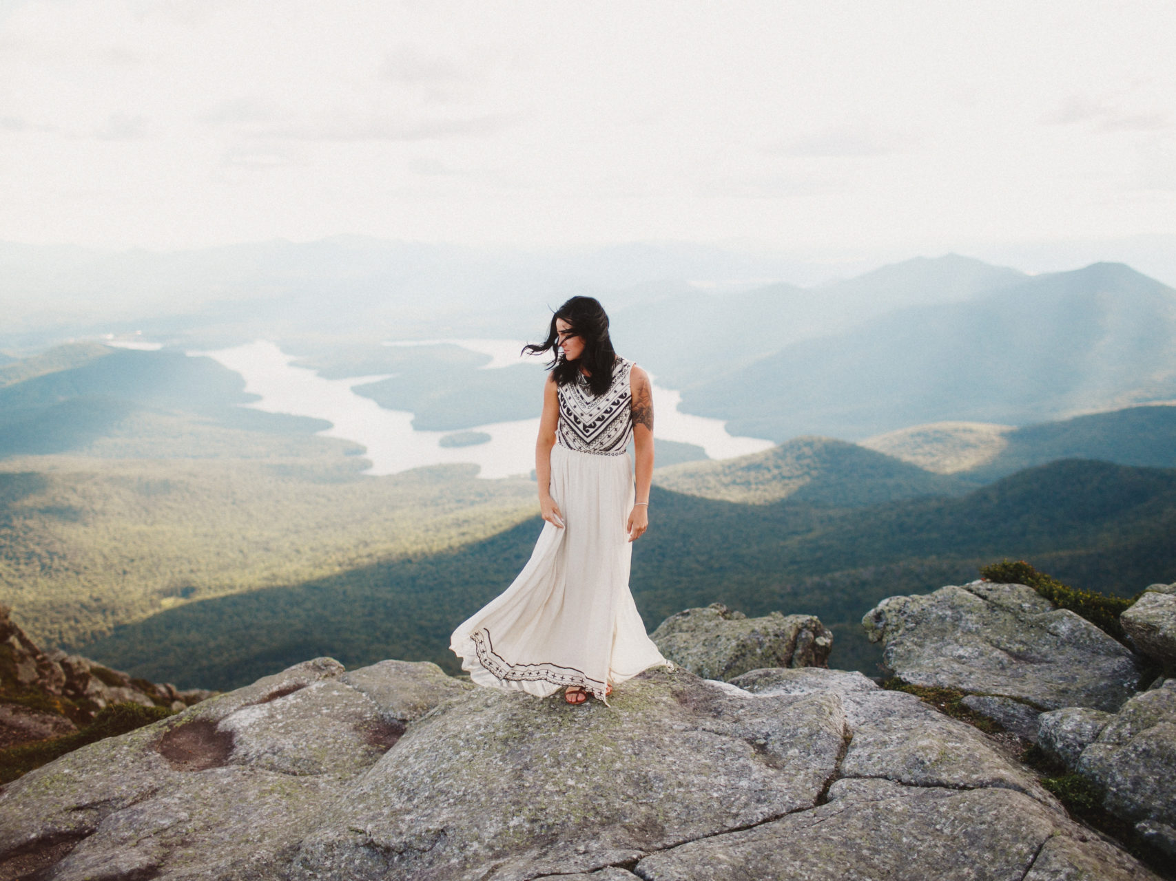 A woman in a flowing white and black gown stands on top of a mountain overlooking a green river valley in a photo by Shaw Photography Co.