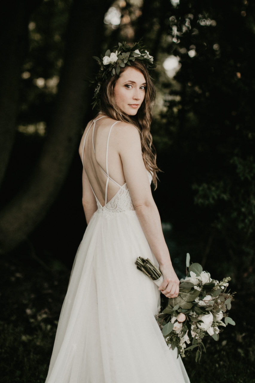 A bride in a long backless wedding dress and flower crown holds a bouquet in her right hand and looks over her shoulder at you in a photo by Shaw Photography Co.