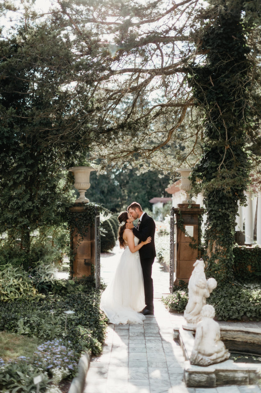 A wedding couple kiss in an ivy covered green garden with white marble cherub statues and urns on top of old stone gate posts in a photo by Shaw Photography Co.