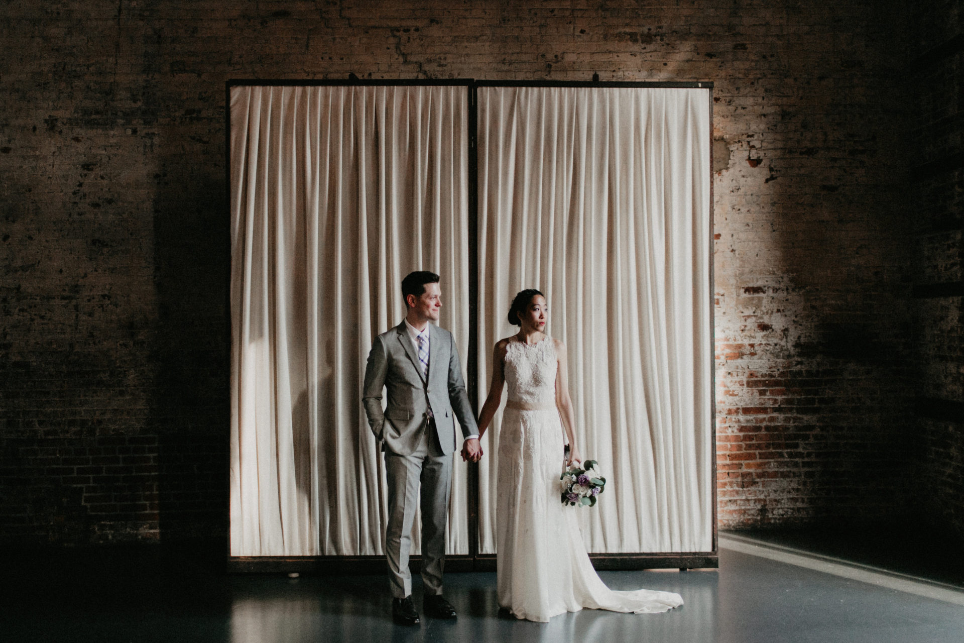A couple in wedding clothes holds hands in front of a freestanding off-white curtain in an industrial space, looking off toward the light in a photo by Shaw Photography Co.
