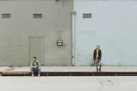 A couple on the street—one sits on the curb and one stands far away from each other, looking in different directions