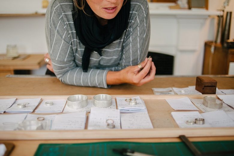wedding rings and other jewelry from ash hilton laid on a table as a woman surveys the colleciton
