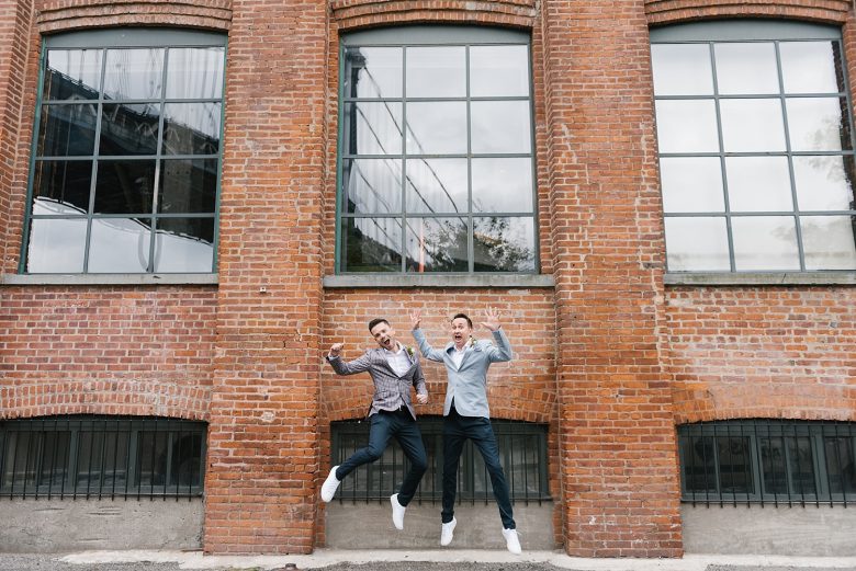 two grooms jumping for joy in front of brick industrial building