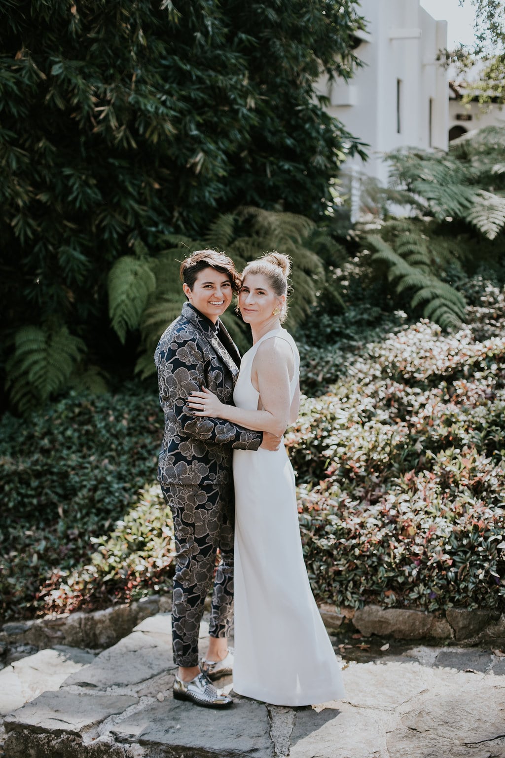 A queer couple in a wedding dress and brocade grey and black suit smile in an embrace in front of botanicals in a Studio XIII Photography image