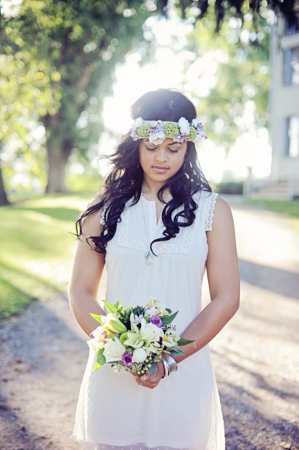 A bride stands in the middle of a long dirt driveway while wearing a flower crown as the sun glimmers through the trees
