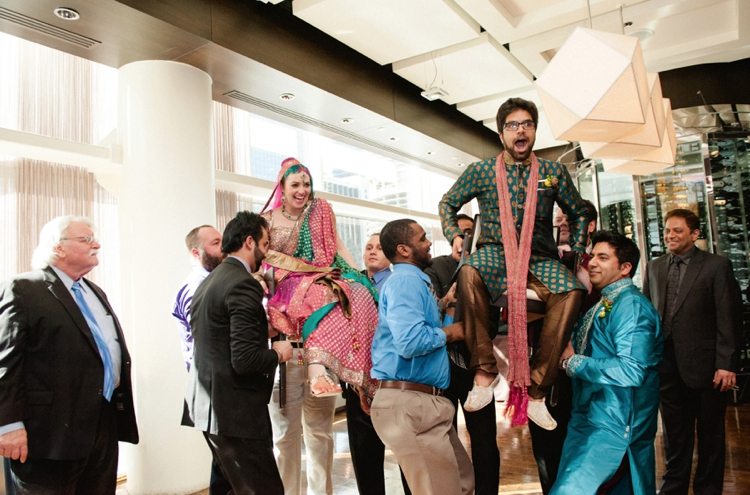 Bride and groom in Indian wedding attire, being lifted in chairs by wedding guests for the Hora