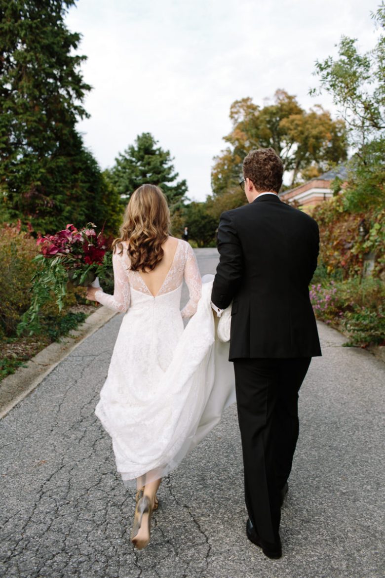 Groom carrying brides train; bride with bouquet; both walking away from camera