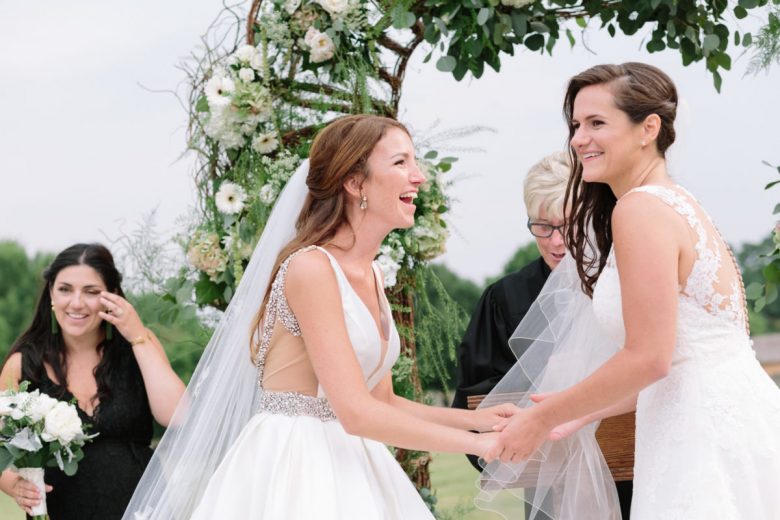 two brides and officiant in front of floral wedding arch, laughing; bridesmaid crying in background