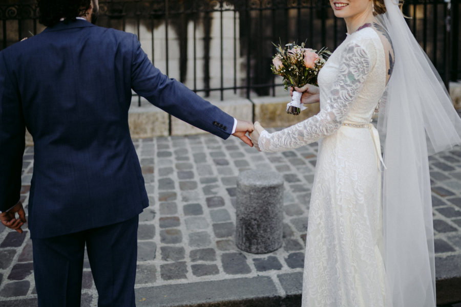 bride in gown with veil, holding bouquet in right hand, holds groom's hand and turns toward you, smiling