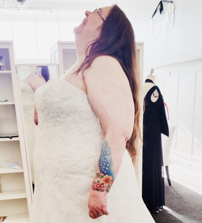 Woman with long hair and tattoo laughing while trying on strapless lace wedding gown in store