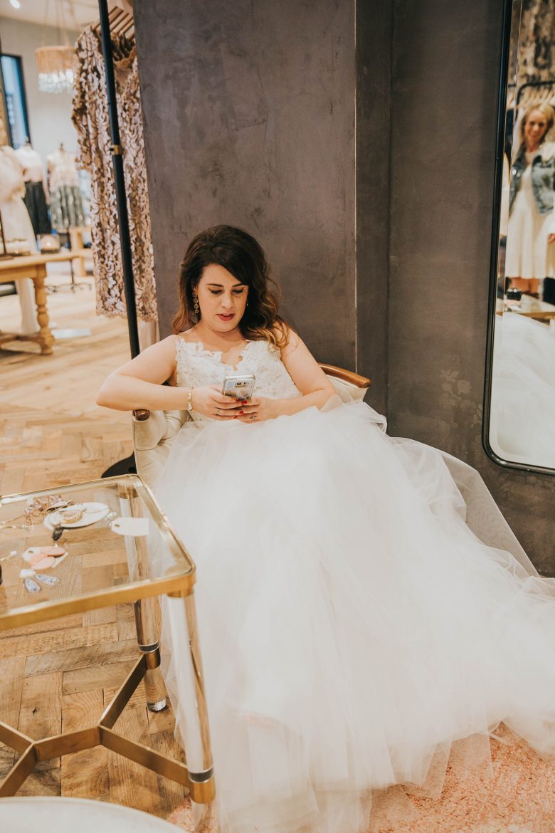 Meg in wedding dress in shop sitting in a chair, checking her phone