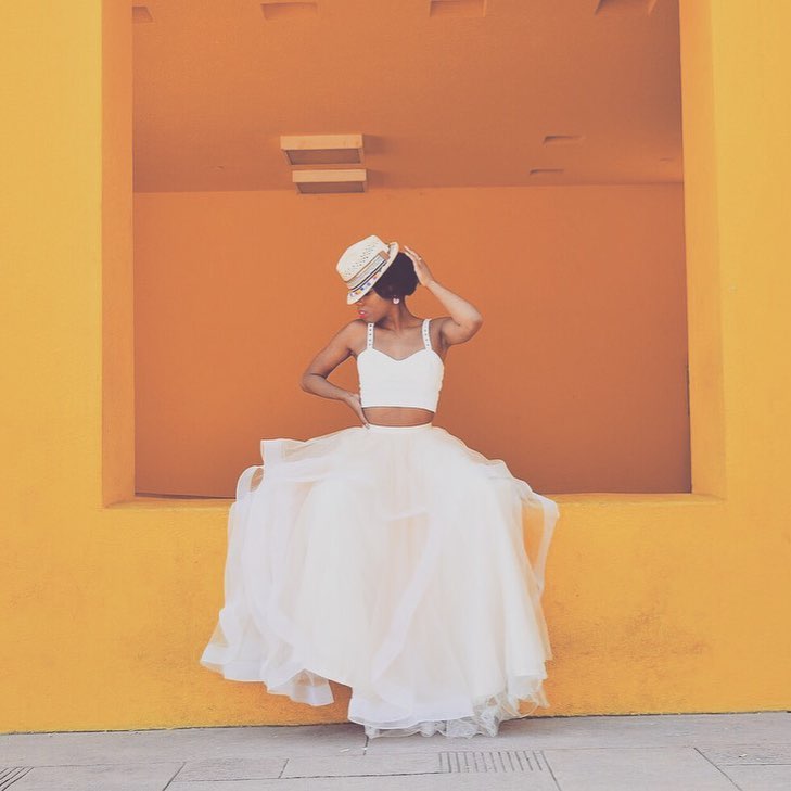 Woman in fedora sitting on yellow and orange wall in crop top wedding dress