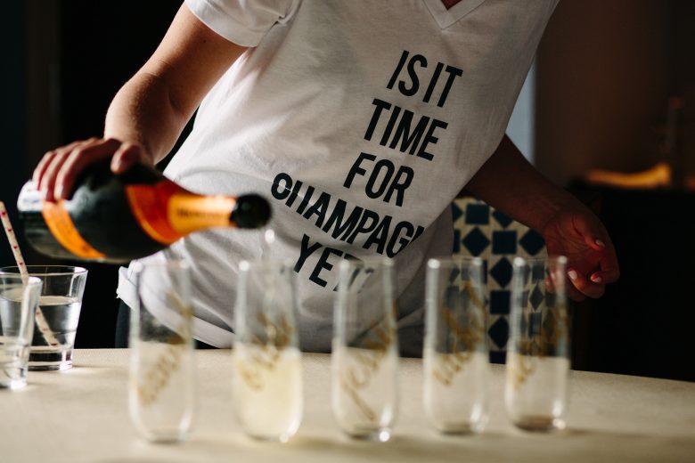 Woman pouring champagne into glasses, wearing a white t-shirt that reads "Is it time for champagne yet?"