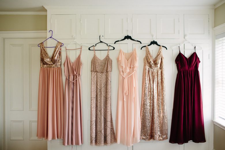 Six full length formal dresses hanging on hangers against a white wall