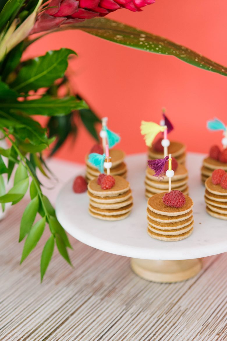 mini pancake stacks with raspberries and toothpicks with tassels on white pedestal platter