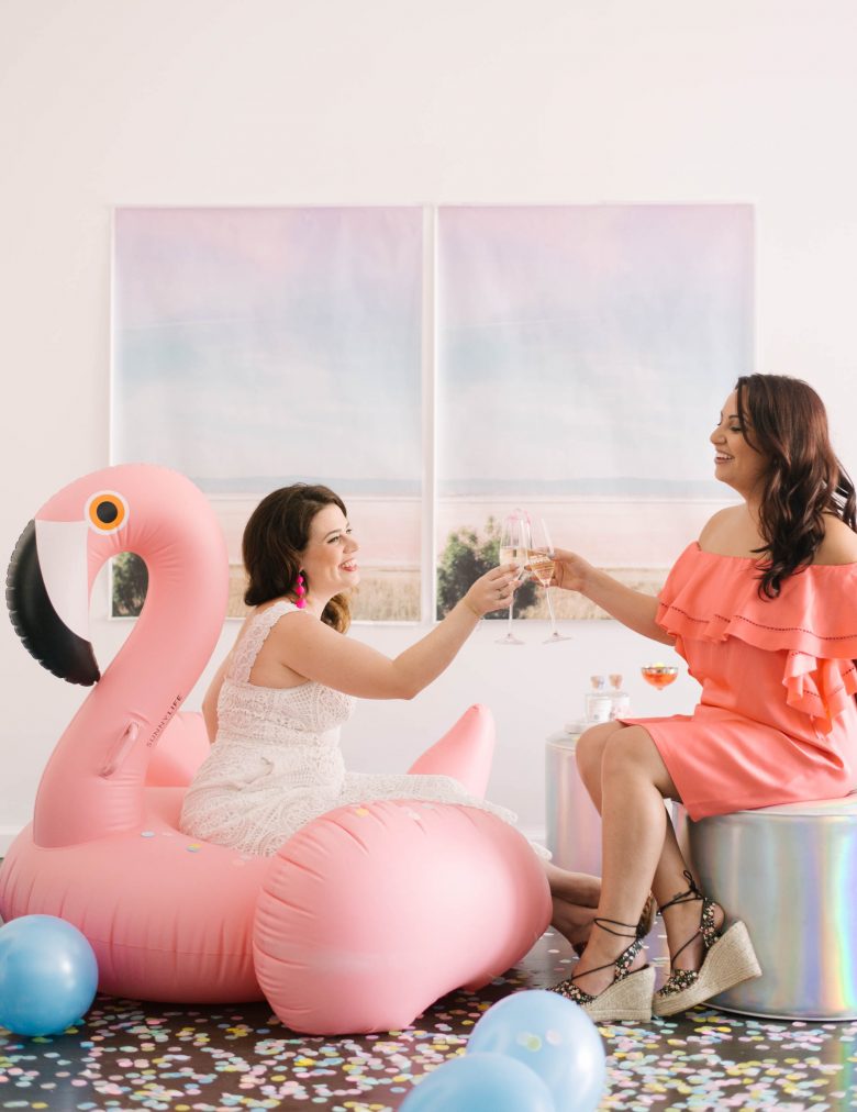 Two women toasting champagne, one on an inflatable pink flamingo, the other on a holographic pouf