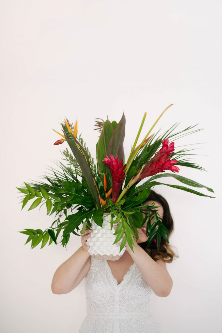 woman in white jumpsuit holding pineapple vase with tropical foliage
