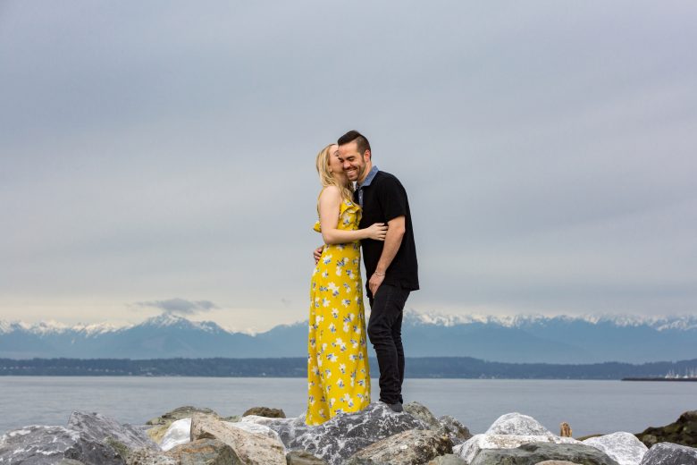 woman in yellow flower dress kissing man in black's cheek as he laughs, as they stand on rocks in front of mountains across water