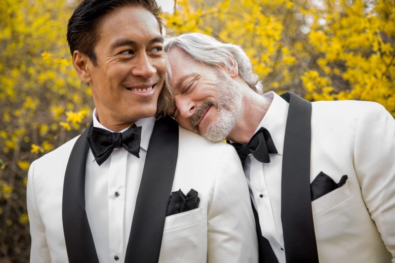 groom with his head on the shoulder of his groom, both in white and black tuxes, with yellow blossoms behind them