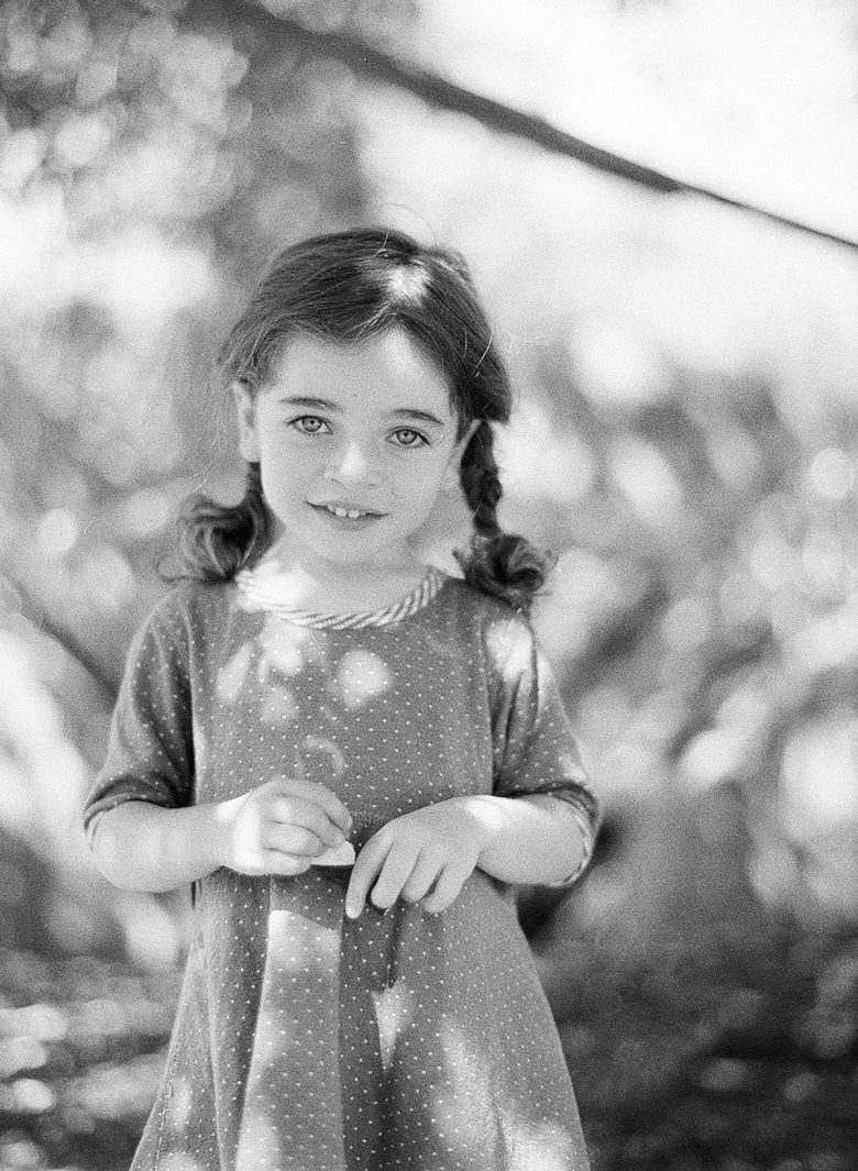 young girl with pigtail braids, smiling, looking at the camera, standing in light filtered through trees, in black and white