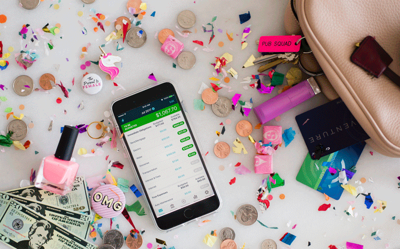 YNAB app on smartphone spilling out of purse with change, confetti, a unicorn pin, a credit card, lipstick, a ring, a pink pin that reads "OMG," pink nail polish, cash, and a keychain that reads "pub squad"