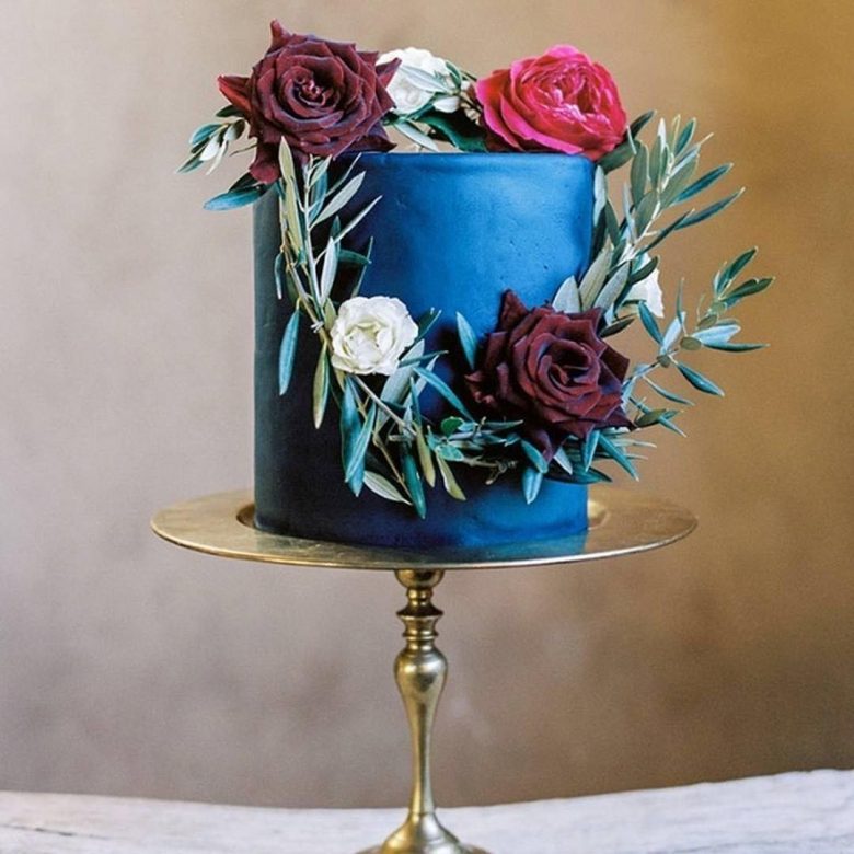 Blue cylindrical cake on gold stand with wreath of roses and greenery on it