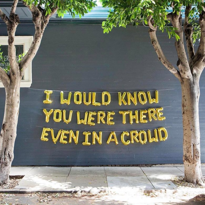 Gold letter balloons strung up on an exterior wall reading "I would know you were there even in a crowd"