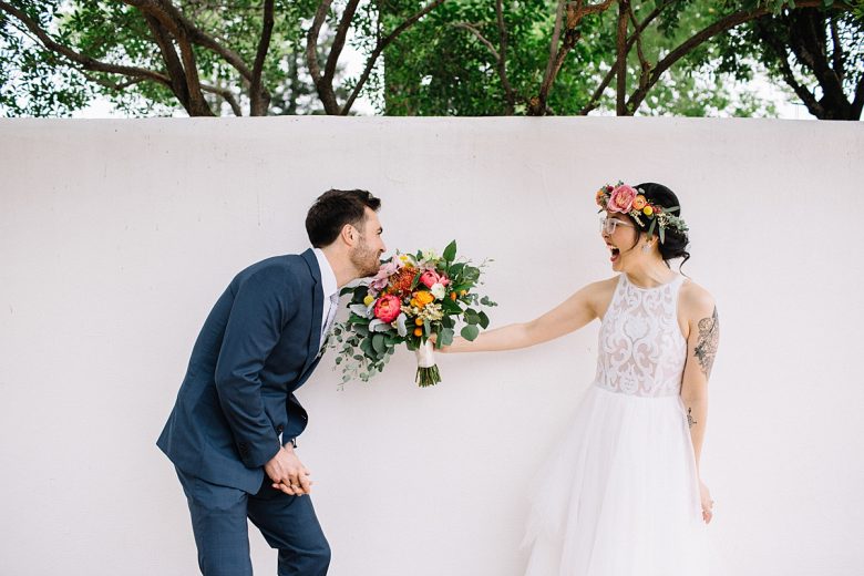 Groom playfully bites at brides bouquet as she laughs