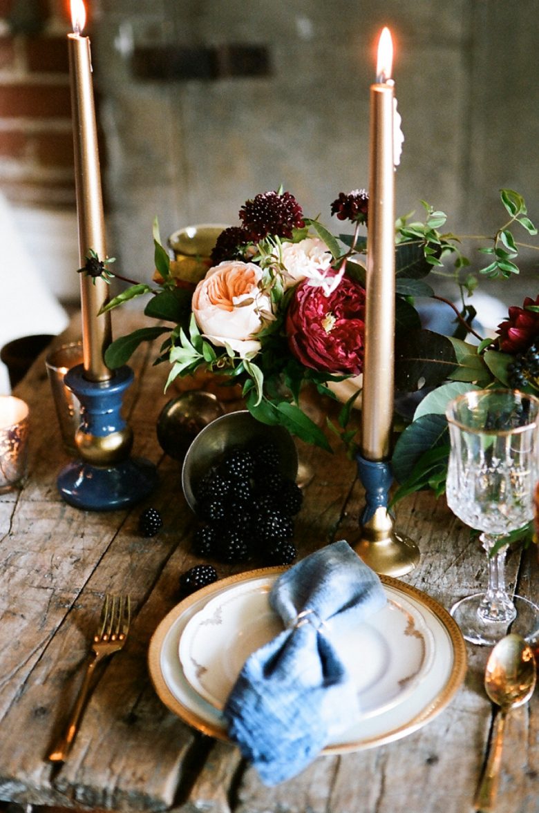 romantic tablescape with gold taper candles, blackberries, dark flowers, gold rimmed plates, on rustic wood table