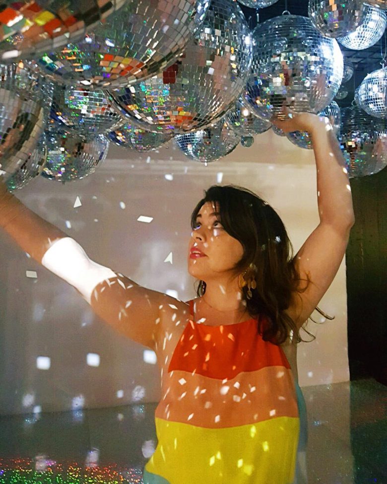 Meg wearing rainbow colored shirt standing under a ceiling filled with disco balls
