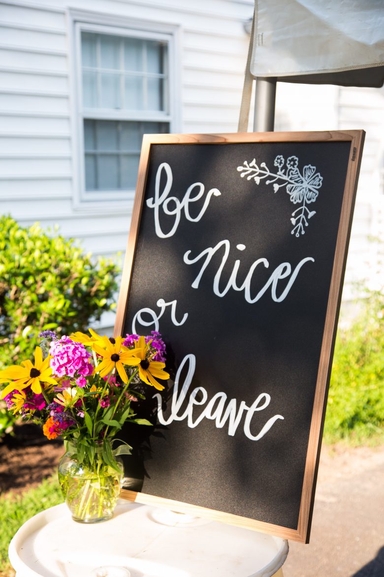 Be nice or leave written on chalkboard sign with drawing of flower next to a floral arrangement on a table near an exterior house wall