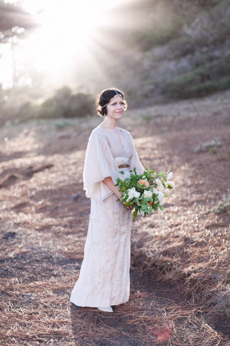 A bride wearing a dress reminiscent of medieval times, stands on a hillside with sunbeams behind her